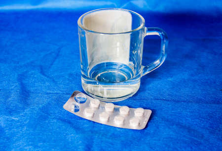 Tablets in a blister with a mug of water next to them. Close-up, blue background.