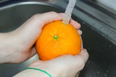 Women's hands wash an orange in the sink under a stream of water. hygiene requirements.の写真素材