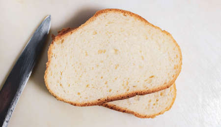 Two cut pieces of a white loaf on a light surface with a knife lying next to it. Top view, close-up.