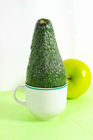 A whole green avocado stands in a mug with an apple in the background. Vertical frame, selective...