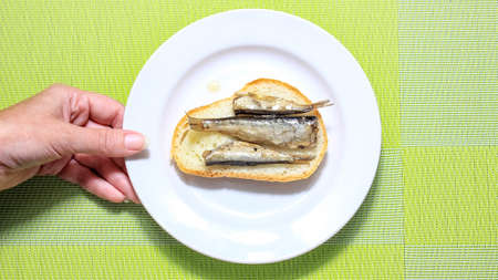 The hand that holds a plate with a sandwich with sprats. Top view.