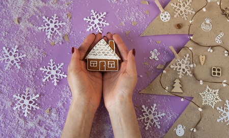 Female hands holding little gingerbread house on table with Christmas decorations. Cut paper in fir-tree shape and snowflakes. View from aboveの写真素材