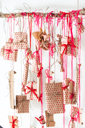 Handmade advent calendar hanging on a white wall. Gifts wrapped in paper and tied with red threads and ribbons. Wooden stick and many presentsの写真素材