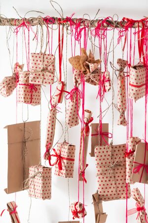 Handmade advent calendar hanging on a white wall. Gifts wrapped in paper and tied with red threads and ribbons. Wooden stick and many presentsの写真素材