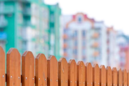 In the foreground is a fence made of planks, in the background are blurry apartment buildings.