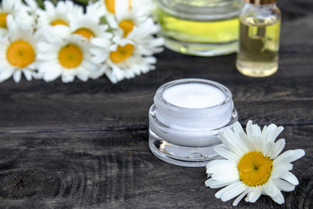 Essential oil and cosmetic cream are on the wooden table near the chamomile flowers.