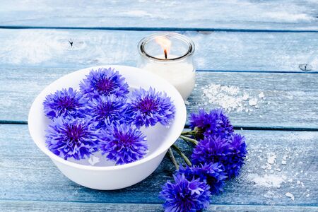 blue cornflowers lie in water in a white bowl near a burning candle against the background of blue old boards. close-upの写真素材