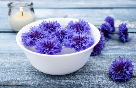 blue cornflowers lie in water in a white bowl near a burning candle against the background of blue old boards. close-upの写真素材