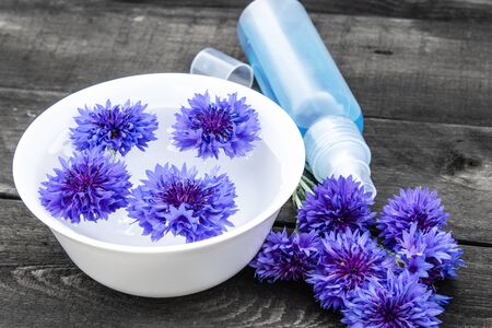 blue cornflowers lie in the water in a bowl near a bottle of cornflower water against the background of old boards. close-upの写真素材