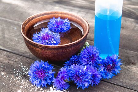 blue cornflowers lie in the water in a bowl near a bottle of cornflower water against the background of old boards. close-upの写真素材