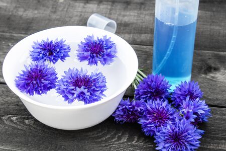 blue cornflowers lie in the water in a bowl near a bottle of cornflower water against the background of old boards. close-upの写真素材