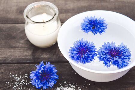 blue cornflowers lie in the water in a white bowl near a burning candle on the background of old boards. close-upの写真素材