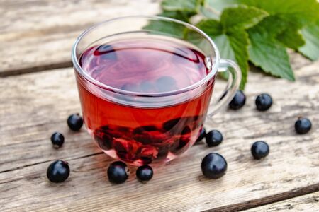 A cup of tea with black currants on a wooden table next to green leaves and currant berries. Close-up.の写真素材