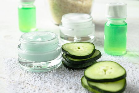Face cream in a glass jar with cucumber extract next to slices of fresh cucumbers on a white wooden background. Spa treatments.の写真素材