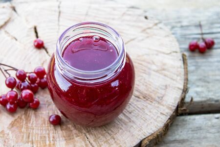 Viburnum fruit jam in a glass jar on a wooden table near the ripe red viburnum berries. Source of...