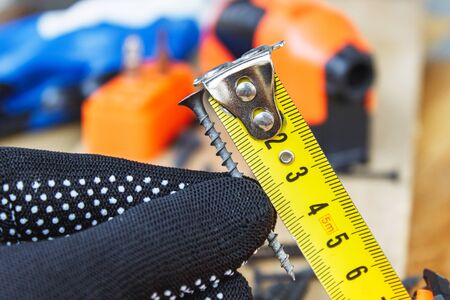 The black screw in hand and is measured by a yellow tape measure with a ruler on a wooden board background. The concept of tools and repair work. Steel screws. Metal screwの写真素材