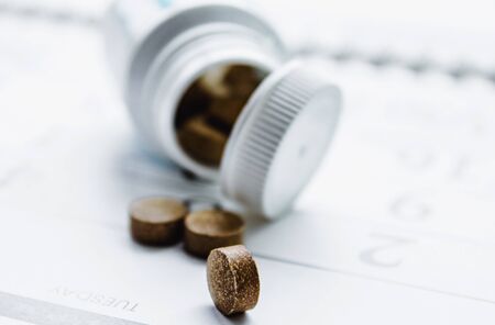 Small dark pills next to a white jar or bottle on a calendar background. The cure for the disease....