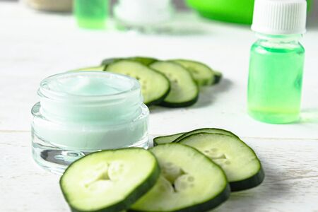 Face cream in a glass jar with cucumber extract next to slices of fresh cucumbers on a white wooden background. Spa treatments.の写真素材