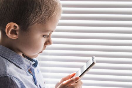 A child by the window with a mobile phone. Home schooling, Social distance. The concept of distance learning and online learning.の写真素材