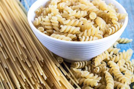 Dry pasta in a bowl on a background of blue boards near a pile of different pasta. Healthy eating or healthy lifestyle.の写真素材
