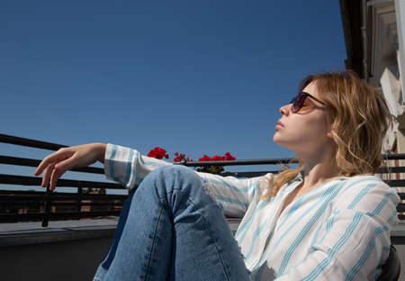 Beautiful girl in sunglasses sits in a chair on the balcony in the morning lightの写真素材
