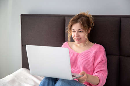 A beautiful girl is sitting in pink sweater with a laptop , chatting.の写真素材