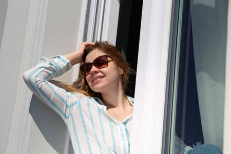 Happy blonde woman in sunglasses sits on windowsill and smiles at the sun