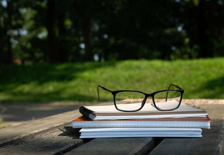 glasses lie on top of a pile of books with a notebook on a bench in the parkの写真素材