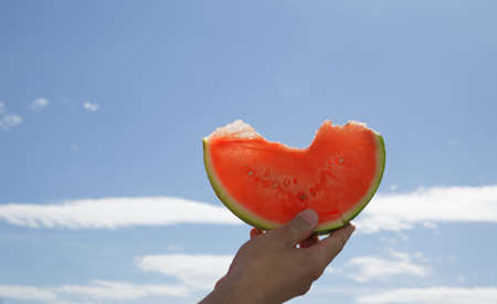 hand holding a slice of watermelon against the blue skyの写真素材