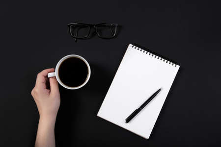 hand holds a cup of coffee on a working black table next to notepadの写真素材