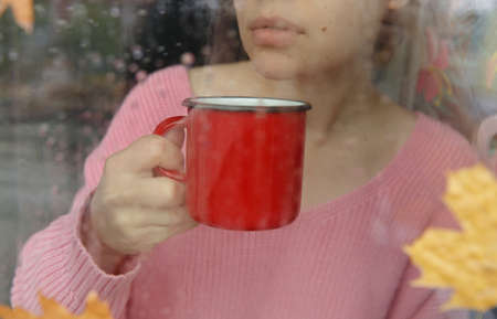 girl with red coffee mug behind wet window with raindrops on glassの写真素材