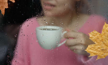 girl with white cup behind wet window with raindrops on glassの写真素材