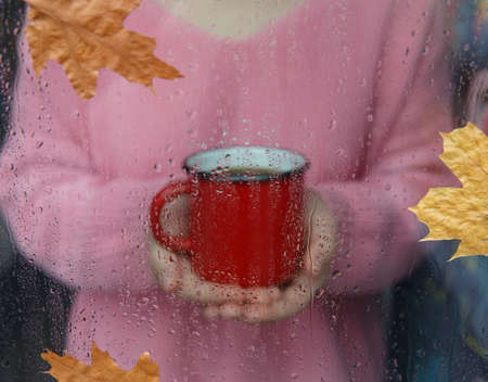 red coffee mug in hands and window with raindropsの写真素材