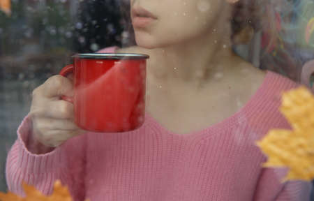 girl holding a red mug with coffee near the window with raindropsの写真素材