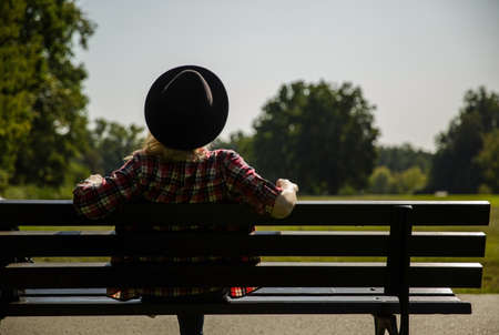 young woman in a hat and a plaid shirt sits on a bench near a meadow in the parの写真素材