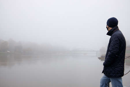 man in mask and hat stands on the bank of the river over which the fog hangs