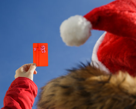 Close-up of a female hand holding a gift card with a picture of sweets against a blue sky. Wearing a Santa hat and a red jacket. gift or discount concept. selective focusの写真素材