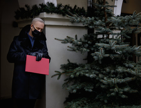 a gray-haired man in a black coat holds a red folder in his hands and watches the clock. the concept of waiting and meeting. High quality photoの写真素材