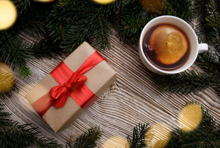 gift box with a red bow and a cup with a fragrant hot drink with fruit stands on a wooden table between fir branches. concept for Christmas and New Year holidays.light bulbs garlands aroundの写真素材