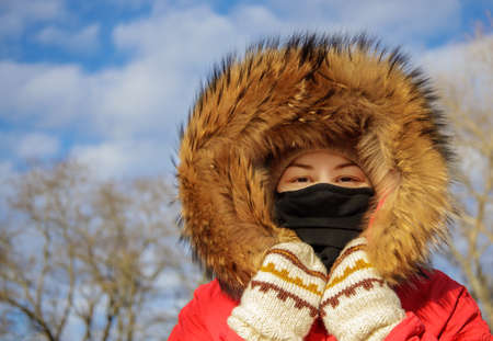 cute girl in a mask and a red jacket with a fur hood and mittens in winter against the background of...