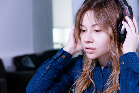 A young woman in a blue blouse holding headphones with her hands while sitting at a table with a laptop and looking at the screen. High quality photoの写真素材