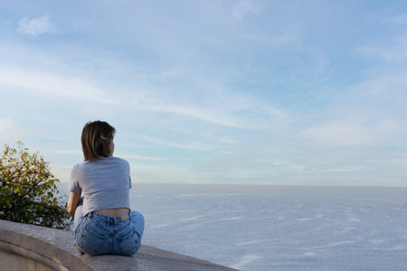 A young woman in jeans and a T-shirt sits on a stone slab and looks at the sea. High quality photoの写真素材