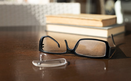 a broken eyeglass frame lies in front of a stack of old books on a brown wooden table. High quality photoの写真素材