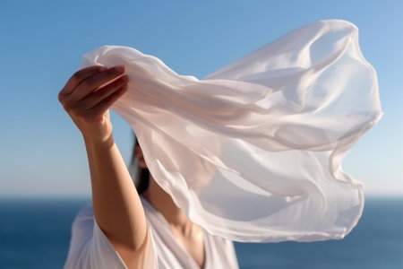 a womans hand holds a light white transparent fabric fluttering in the wind against the backdrop of the sea on a sunny dayの素材