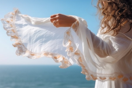 a womans hand holds a light white transparent fabric fluttering in the wind against the backdrop of the sea on a sunny dayの素材