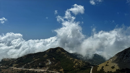 A captivating view of clouds rolling over a lush mountain range, with a winding road cutting through...