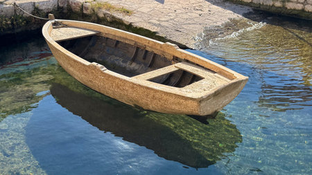 Wooden rowboat tied with rope on calm clear water at small harbor, reflecting environment and sunlight.の写真素材