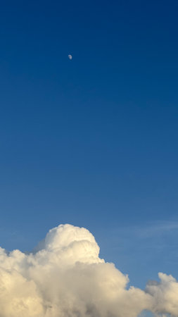 Minimalistic composition of crescent moon in deep blue sky above fluffy white clouds illuminated by...