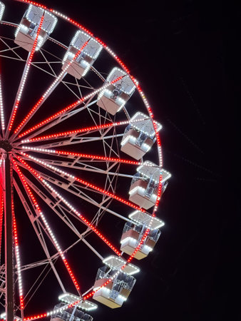 A close-up of a Ferris Wheel illuminated with red and white lights against a dark night skyの写真素材