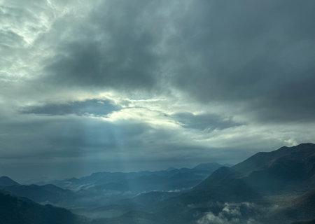A dramatic sky with thick clouds allowing beams of sunlight to stream through, illuminating the misty mountain landscape below.の写真素材